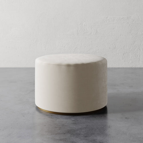 Lemieux et Cie Sophie Round Ottoman in ivory upholstery with brass base detail, cylindrical ottoman on concrete floor against textured plaster wall.