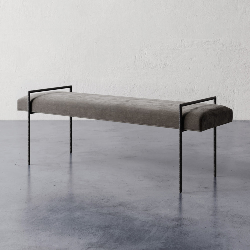 Lemieux et Cie Imane 60" Bench in charcoal upholstery with hand-hammered bronze frame, sculptural bench shown on concrete floor against textured white plaster wall.