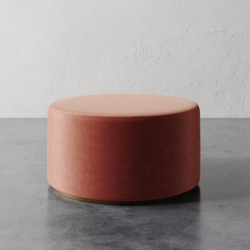 Lemieux et Cie Sophie Round 36" Ottoman in warm blush upholstery with brass base, low-profile ottoman on polished concrete floor against textured plaster walls.
