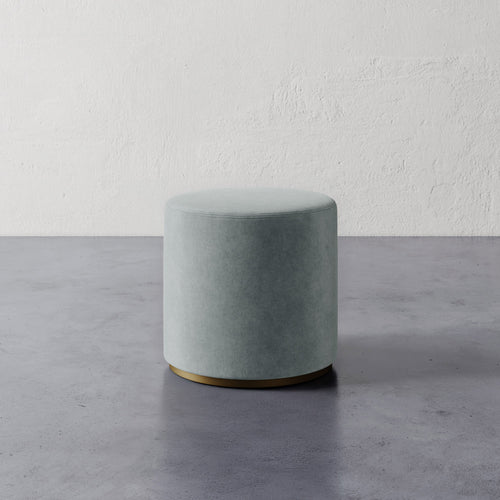Lemieux et Cie Sophie Round Ottoman in soft blue-gray upholstery with brass base, cylindrical ottoman on concrete floor against textured plaster walls.