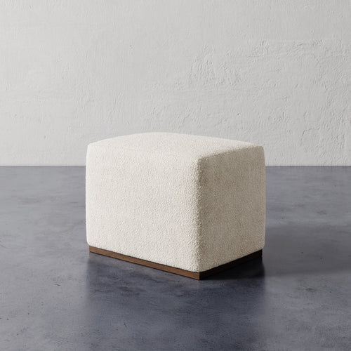 Lemieux et Cie Sophie Rectangular Ottoman in ivory bouclé with wood base, compact ottoman on concrete floor against textured plaster wall.