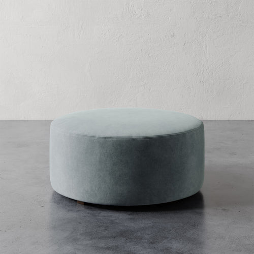 Lemieux et Cie Fabien 36" Round Ottoman in soft sage upholstery, low-profile circular ottoman on concrete floor against textured plaster wall.
