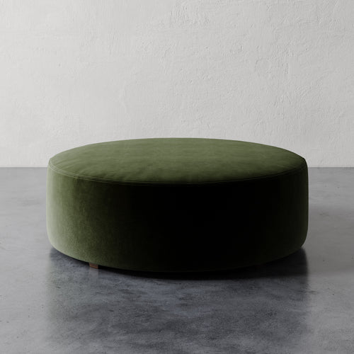 Lemieux et Cie Fabien 48" Round Ottoman in olive green, low-profile ottoman on stone floor against textured plaster walls.
