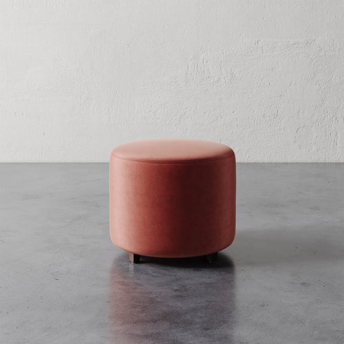 Lemieux et Cie Fabien 22" Round Ottoman in terracotta upholstery, cylindrical ottoman on polished concrete floor against textured white plaster wall.
