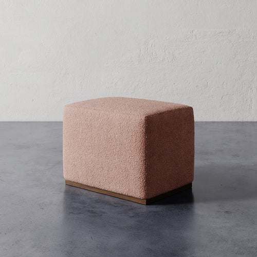 Lemieux et Cie Sophie Rectangular Ottoman in blush bouclé upholstery with dark wood base, shown on concrete floor against textured plaster walls.