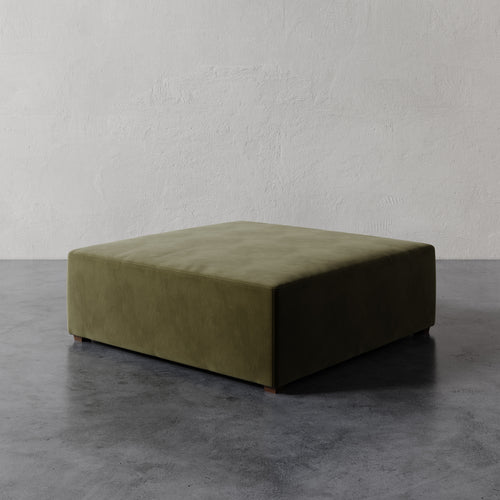 Lemieux et Cie Fabien 48" Square Ottoman in olive green, low-profile upholstered ottoman on stone floor against textured plaster wall.
