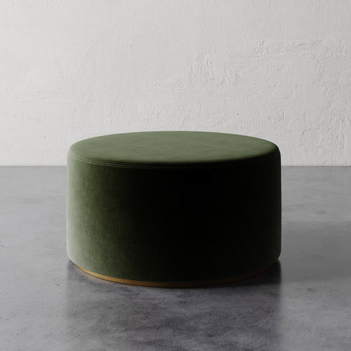 Lemieux et Cie Sophie Round Ottoman in olive green upholstery with brass base detail, low-profile seating on gray concrete floor against textured white plaster wall.