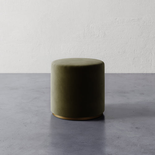 Lemieux et Cie Sophie Round Ottoman in moss green velvet with brass base, cylindrical ottoman on concrete floor against textured plaster walls.