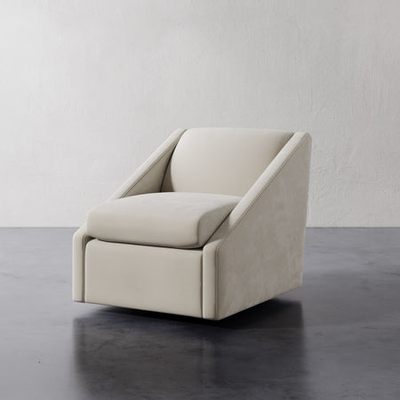 Lemieux et Cie Fontaine Swivel Chair in ivory upholstery, sculptural lounge chair with angled arms on dark stone floor against textured plaster wall.