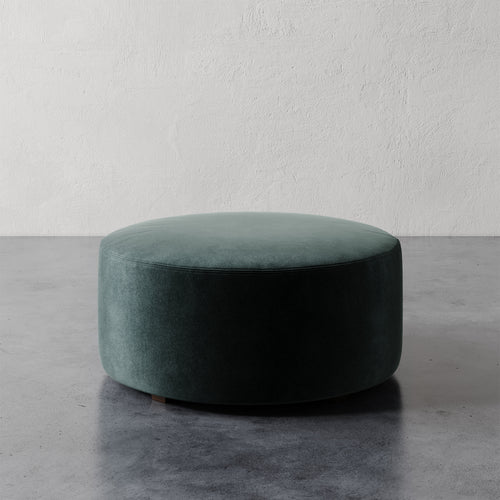 Lemieux et Cie Fabien 36" Round Ottoman, cylindrical upholstered ottoman in deep teal, on polished concrete floor against textured plaster wall.