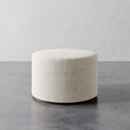 Lemieux et Cie Sophie Round Ottoman in ivory bouclé, low cylindrical ottoman on polished wood base, shown on concrete floor against textured plaster wall.