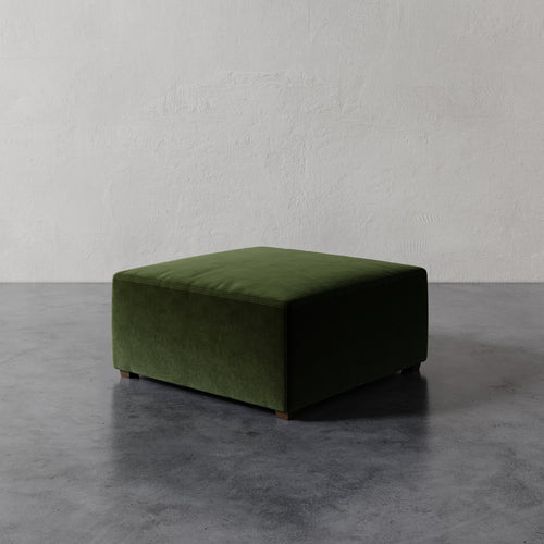 Lemieux et Cie Fabien 36" Square Ottoman in deep olive green, low-profile upholstered ottoman on dark concrete flooring against textured plaster walls.