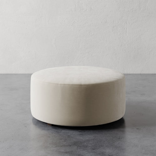 Lemieux et Cie Fabien 36" Round Ottoman in ivory upholstery, low-profile cylindrical ottoman on concrete floor against textured plaster wall.
