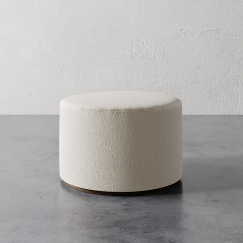 Lemieux et Cie Sophie Round Ottoman in ivory upholstery, cylindrical ottoman with curved base, shown on concrete floor against textured plaster wall.