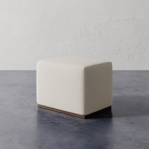 Lemieux et Cie Sophie Rectangular Ottoman in neutral upholstery with wood base, compact ottoman on concrete floor against textured plaster walls.