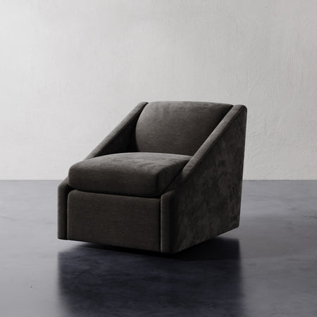 Lemieux et Cie Fontaine Swivel Chair in charcoal upholstery, sculptural lounge chair with angular arms, shown on dark stone floor against white plaster wall.