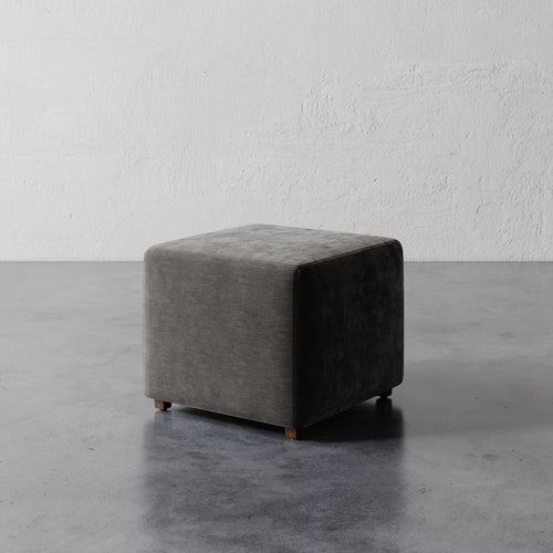 Lemieux et Cie Fabien 22" Square Ottoman in charcoal upholstery, cube-form ottoman on tapered legs, shown on concrete floor against textured plaster walls.