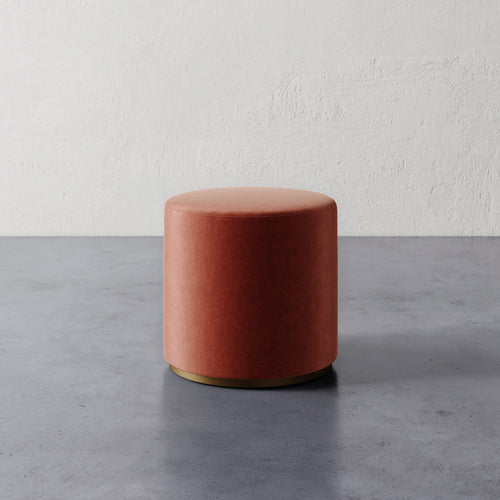 Lemieux et Cie Sophie Round Ottoman in terracotta velvet with brass base, cylindrical ottoman on polished concrete floor against textured plaster wall.