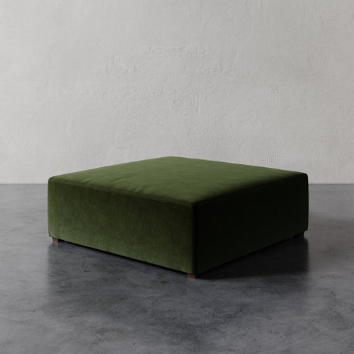 Lemieux et Cie Fabien 48" Square Ottoman in deep olive green, low-profile upholstered ottoman on dark concrete floor against textured plaster wall.