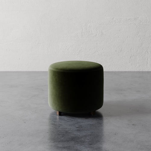 Lemieux et Cie Fabien 22" Round Ottoman in deep olive green upholstery, low cylindrical ottoman on stone floor against textured plaster wall.