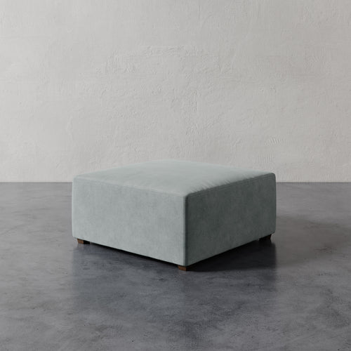Lemieux et Cie Fabien 36" Square Ottoman in soft sage upholstery, low-profile ottoman on dark concrete floor against textured plaster wall.