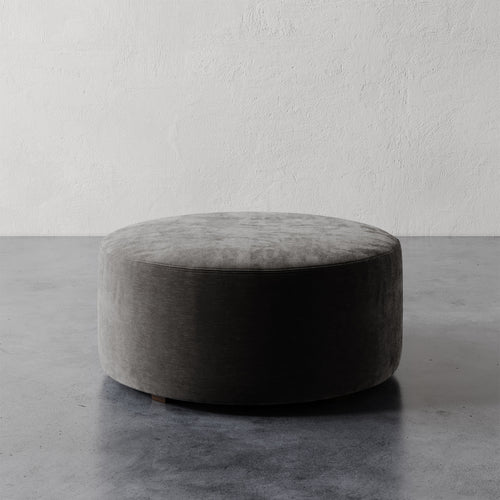 Lemieux et Cie Fabien 36" Round Ottoman, low-profile upholstered ottoman in gray velvet, shown on concrete floor against textured plaster wall.