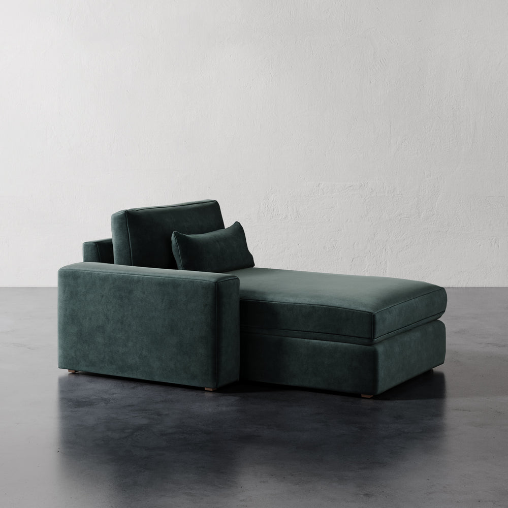 Lemieux et Cie Lacroix Left or Right Chaise & A Half in deep green velvet, modular sectional piece on dark concrete floors against textured white walls.