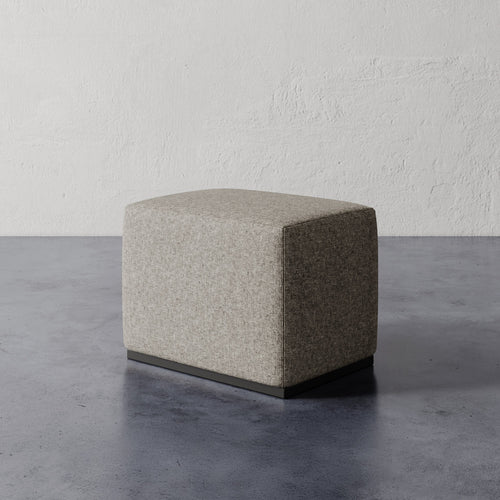 Lemieux et Cie Sophie Rectangular Ottoman in warm neutral upholstery with dark base, compact rectangular ottoman on concrete floor against textured plaster walls.
