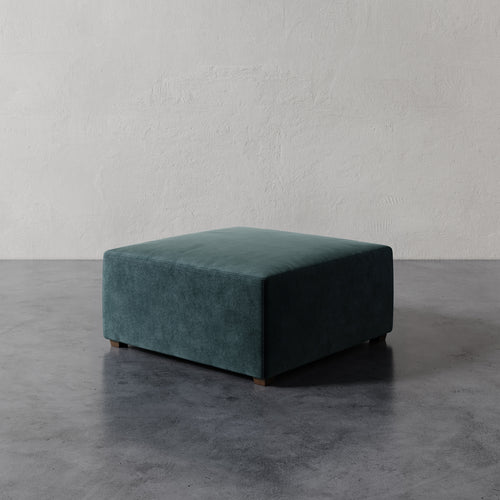 Lemieux et Cie Fabien 36" Square Ottoman in deep teal upholstery, low-profile square ottoman on wood legs, shown on concrete floor against textured plaster wall.