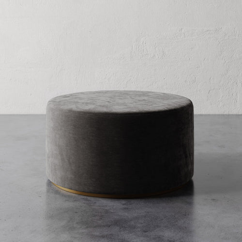 Lemieux et Cie Sophie Round Ottoman in charcoal upholstery with brass base detail, low-profile ottoman on concrete floor against textured plaster wall.