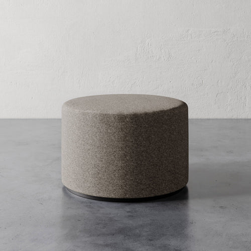 Lemieux et Cie Sophie Round Ottoman in warm grey upholstery, cylindrical ottoman on dark base, shown on concrete floor against textured plaster walls.
