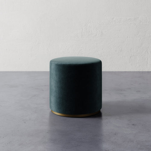Lemieux et Cie Sophie Round Ottoman in deep teal upholstery with brass base, cylindrical ottoman on polished concrete floor against textured plaster wall.