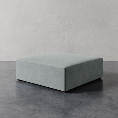 Lemieux et Cie Fabien 48" Square Ottoman in soft sage, low-profile square ottoman on dark concrete floor against textured plaster walls.