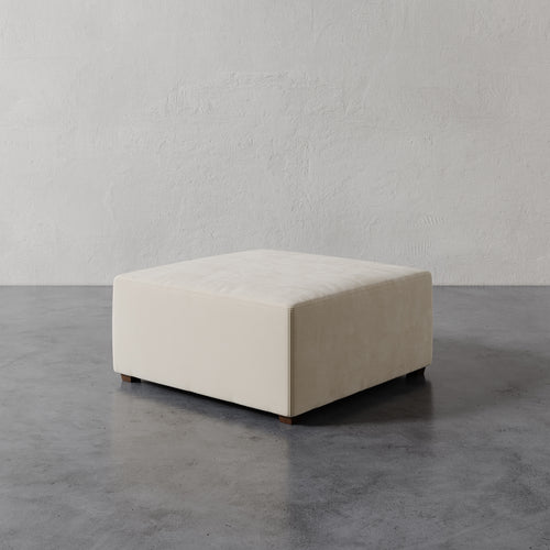 Lemieux et Cie Fabien 36" Square Ottoman in ivory upholstery, low-profile ottoman with tailored seams, shown on concrete floor against textured plaster walls.
