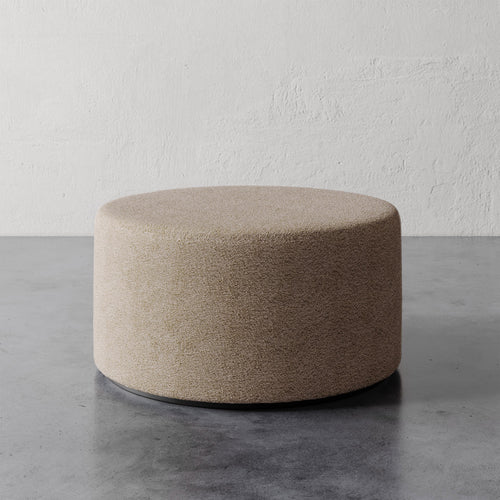 Lemieux et Cie Sophie Round Ottoman in taupe upholstery with dark base, low-profile cylindrical ottoman on concrete floor against textured plaster wall.