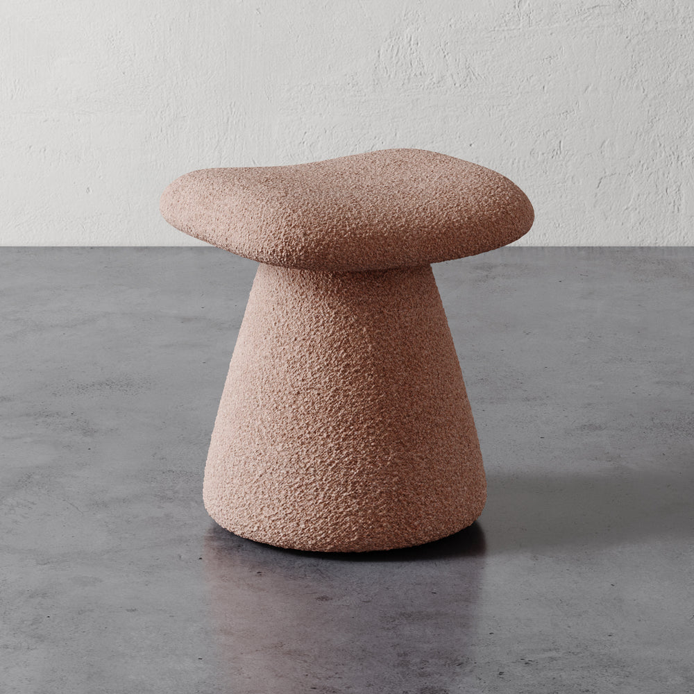 Lemieux et Cie Romy Ottoman in blush textured upholstery, sculptural mushroom-shaped stool with flared pedestal base, shown on concrete floor against textured plaster wall.