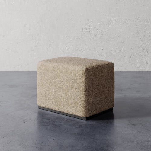 Lemieux et Cie Sophie Rectangular Ottoman in textured neutral upholstery with dark base, compact seating piece on concrete floor against plaster walls.