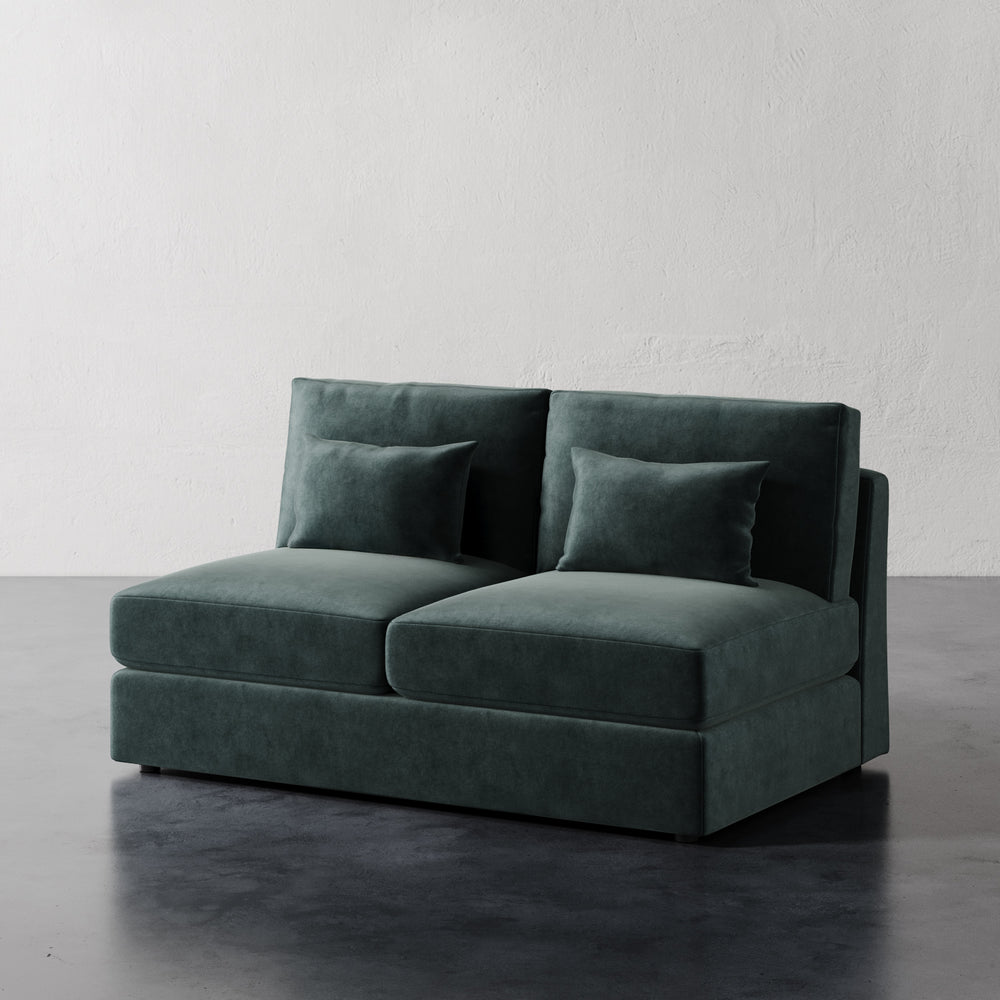 Lemieux et Cie Lacroix Armless Loveseat in deep green velvet, modular sofa with bolster pillows, shown on dark concrete floor against textured plaster wall.