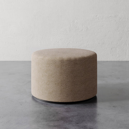 Lemieux et Cie Sophie Round Ottoman in warm taupe upholstery, cylindrical form with dark base, shown on concrete floor against textured plaster wall.