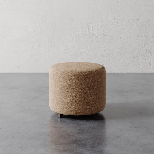 Lemieux et Cie Fabien 22" Round Ottoman in warm bouclé, cylindrical upholstered ottoman on polished concrete floor against textured plaster wall.