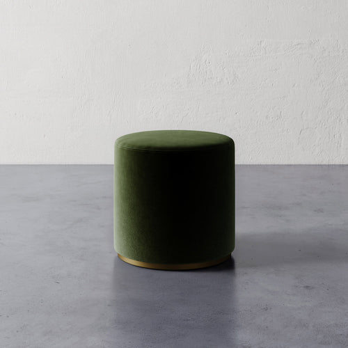 Lemieux et Cie Sophie Round Ottoman in deep green upholstery with brass base, cylindrical ottoman on gray concrete floor against textured plaster walls.