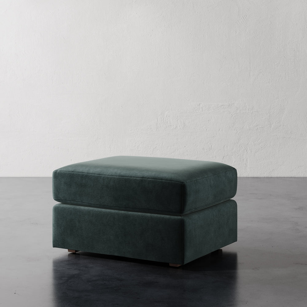 Lemieux et Cie Lacroix Ottoman in deep teal velvet, low-profile upholstered ottoman with rectilinear form, on dark concrete floor against textured white plaster walls.