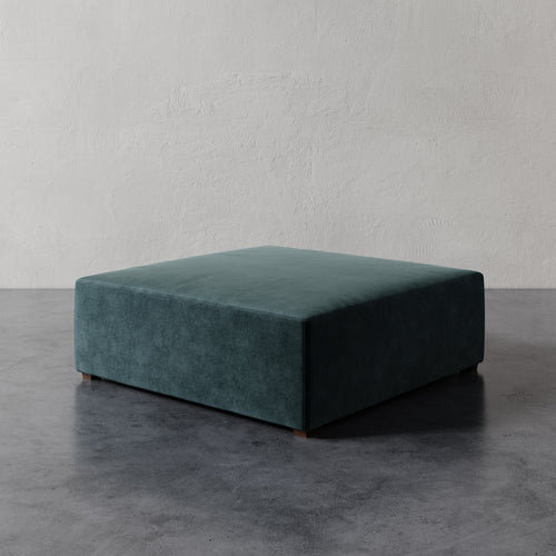 Lemieux et Cie Fabien 48" Square Ottoman in deep teal, low-profile square ottoman on dark concrete floor against textured plaster wall.