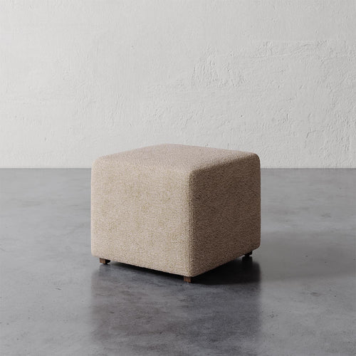 Lemieux et Cie Fabien 22" Square Ottoman, upholstered cube-form ottoman on low legs, shown on concrete floor against textured plaster wall.