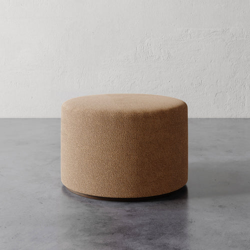 Lemieux et Cie Sophie Round Ottoman in warm tan textured upholstery, cylindrical form on concrete floor against textured plaster wall.