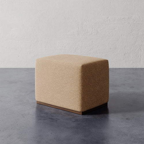 Lemieux et Cie Sophie Rectangular Ottoman in warm textured upholstery with base, shown on concrete floor against plaster wall in soft light.