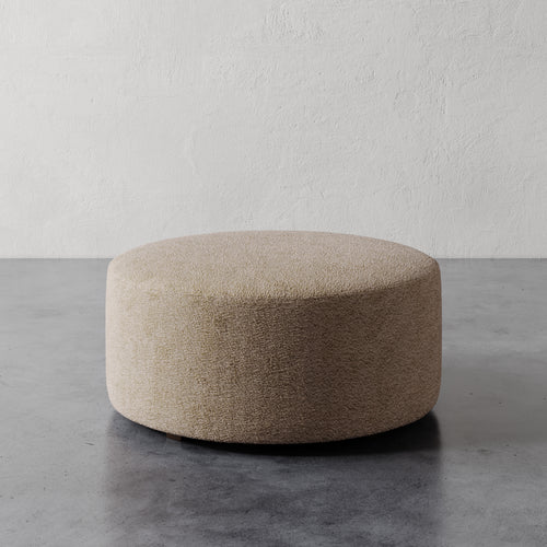 Lemieux et Cie Fabien 36" Round Ottoman in neutral textured upholstery, low-profile round ottoman on concrete floor against textured white plaster wall.