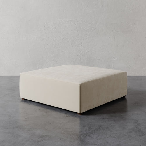 Lemieux et Cie Fabien 48" Square Ottoman in ivory, low-profile square ottoman on stone floor against textured plaster wall.