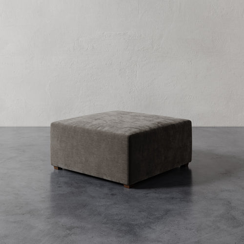 Lemieux et Cie Fabien 36" Square Ottoman in warm gray upholstery, low-profile ottoman on dark concrete floor against textured plaster walls.