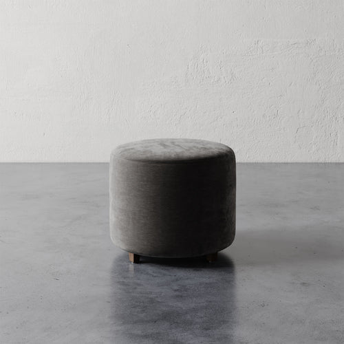 Lemieux et Cie Fabien 22" Round Ottoman, upholstered round ottoman on low feet, charcoal velvet, shown on concrete floor against textured plaster wall.